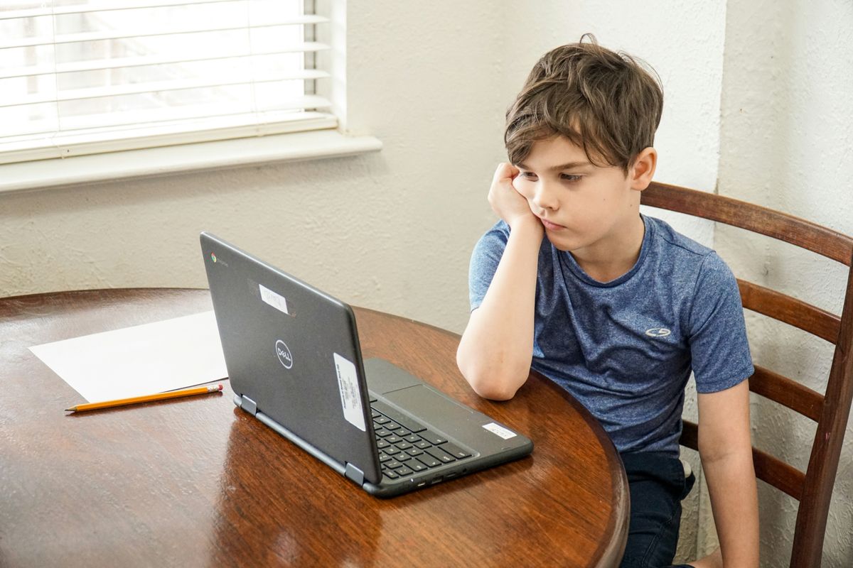 A boy resting his chin on his hand, looking unenthusiastically at a laptop during an online class at home
