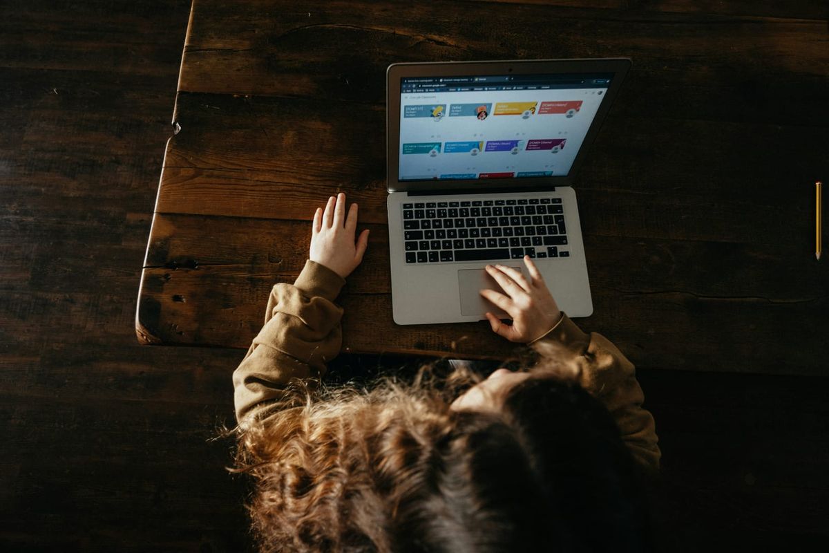 A child seen from above, working on a laptop at a wooden table with one hand on the trackpad