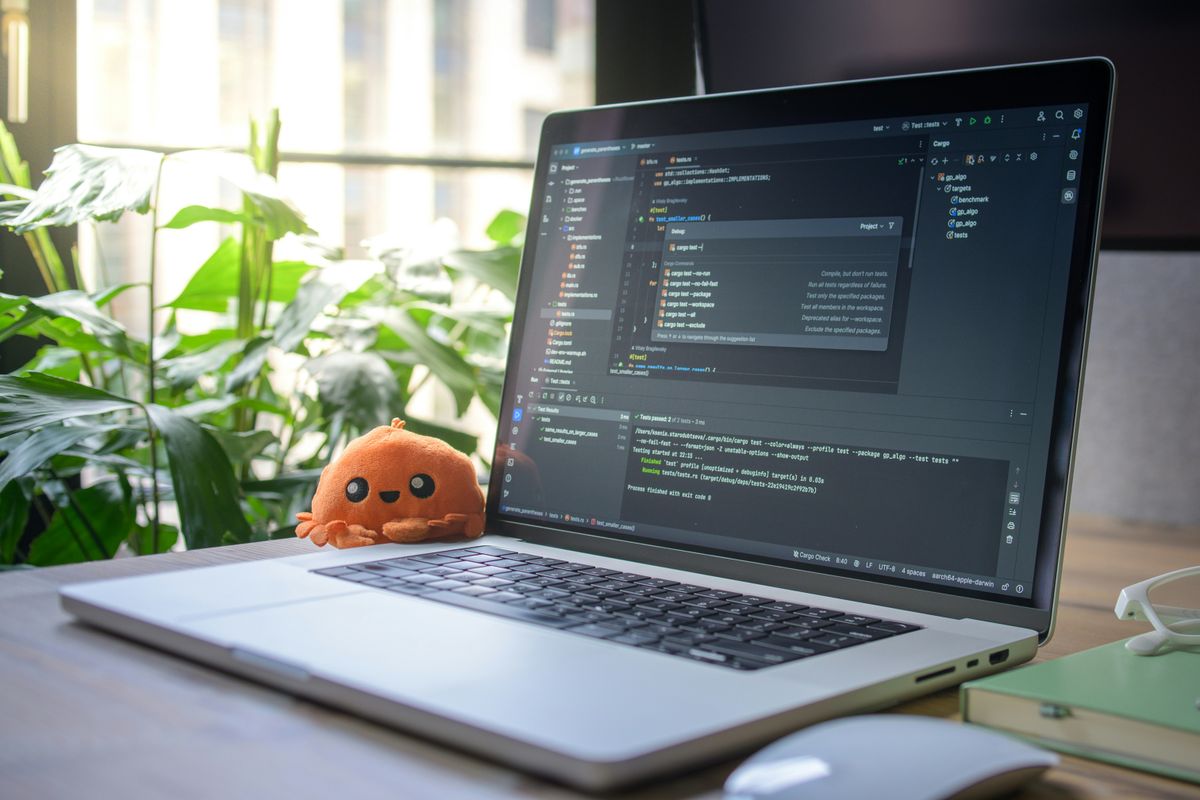 A laptop on a desk showing code and passing tests in a code editor, with a small orange plush toy beside it