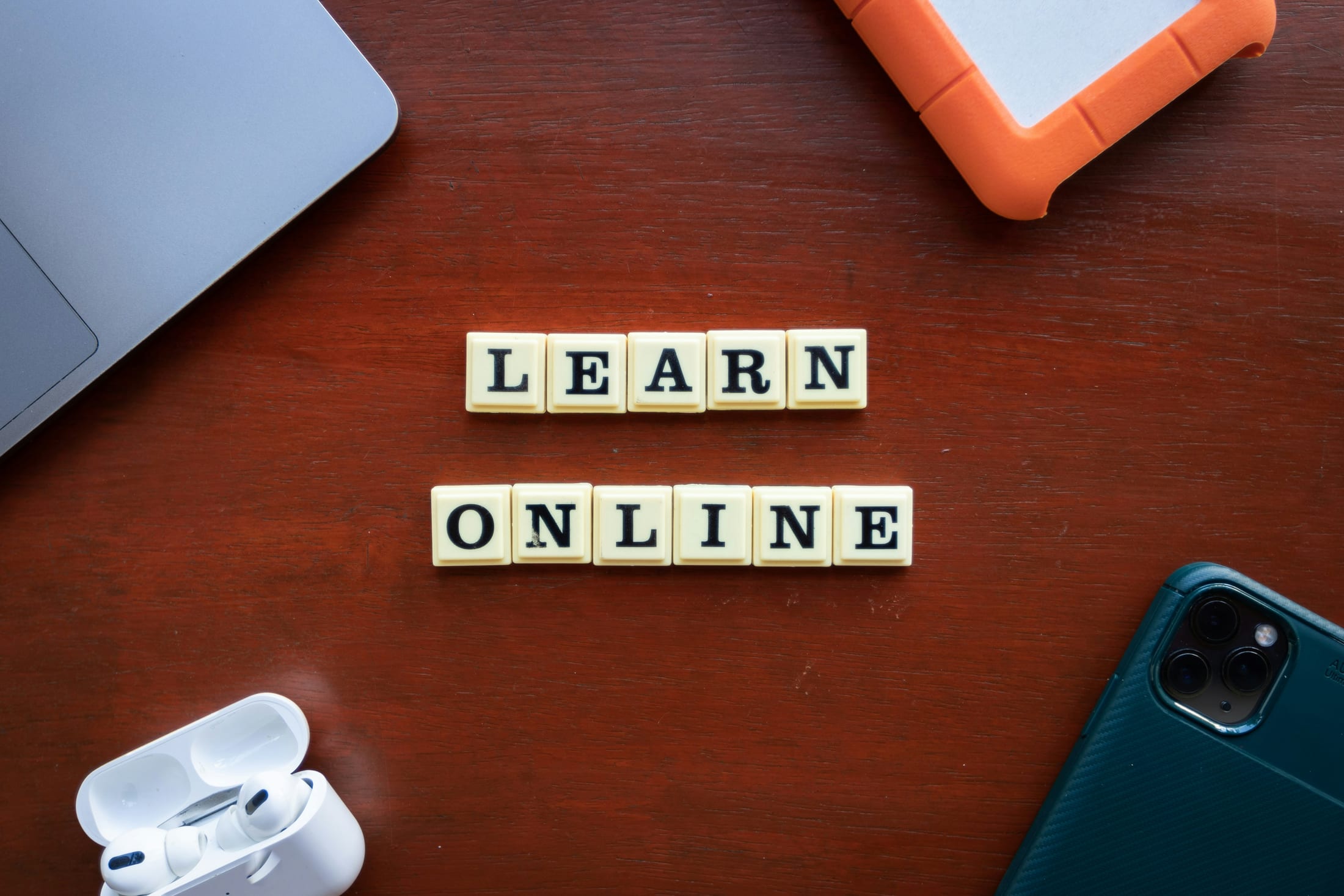 Letter tiles spelling LEARN ONLINE on a desk beside a laptop, phone and earbuds